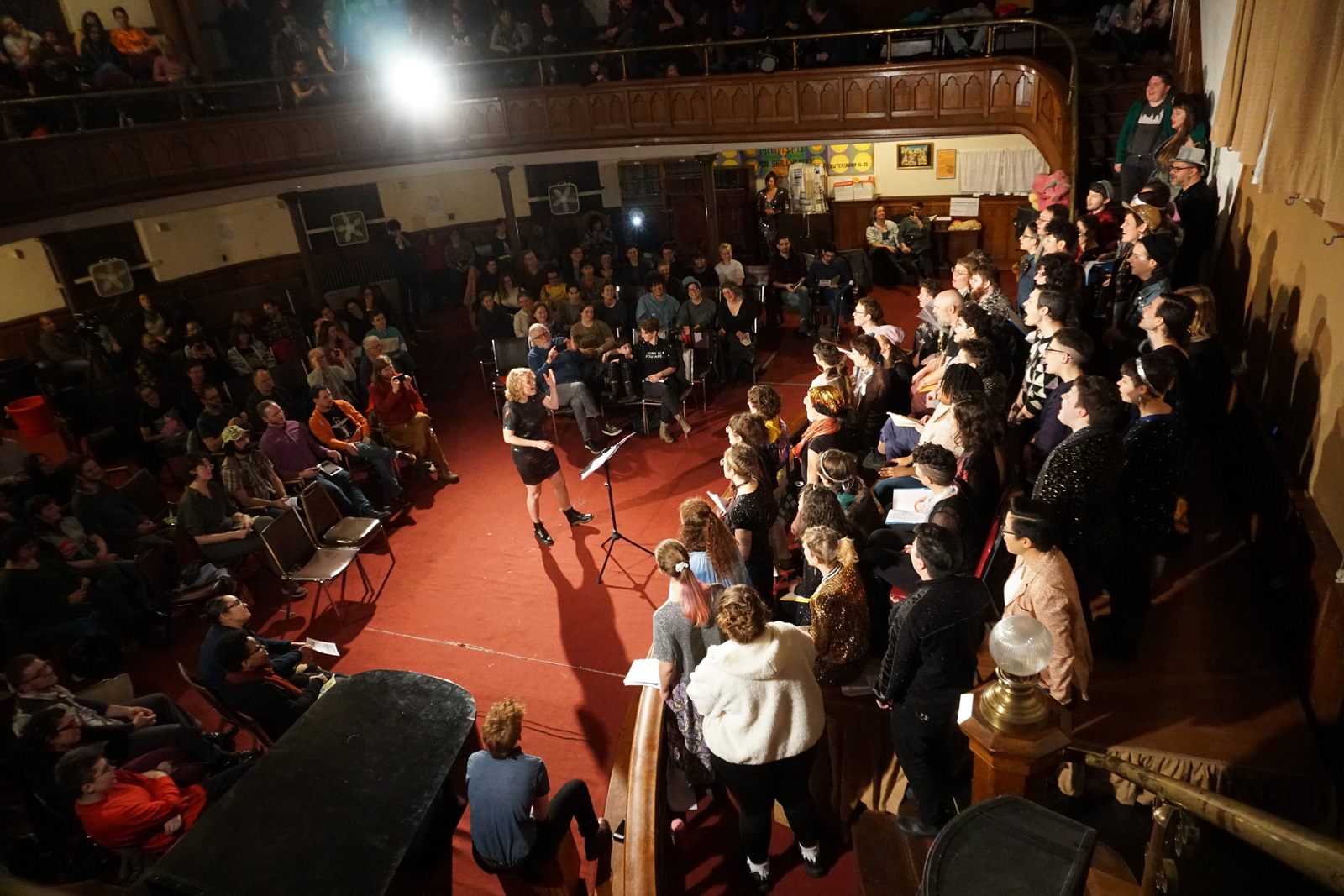 A photograph of a brightly-attired choir performing on a tiered stage before an audience. Emily Bate is energetically conducting, framed on a disk of red carpet in the center.