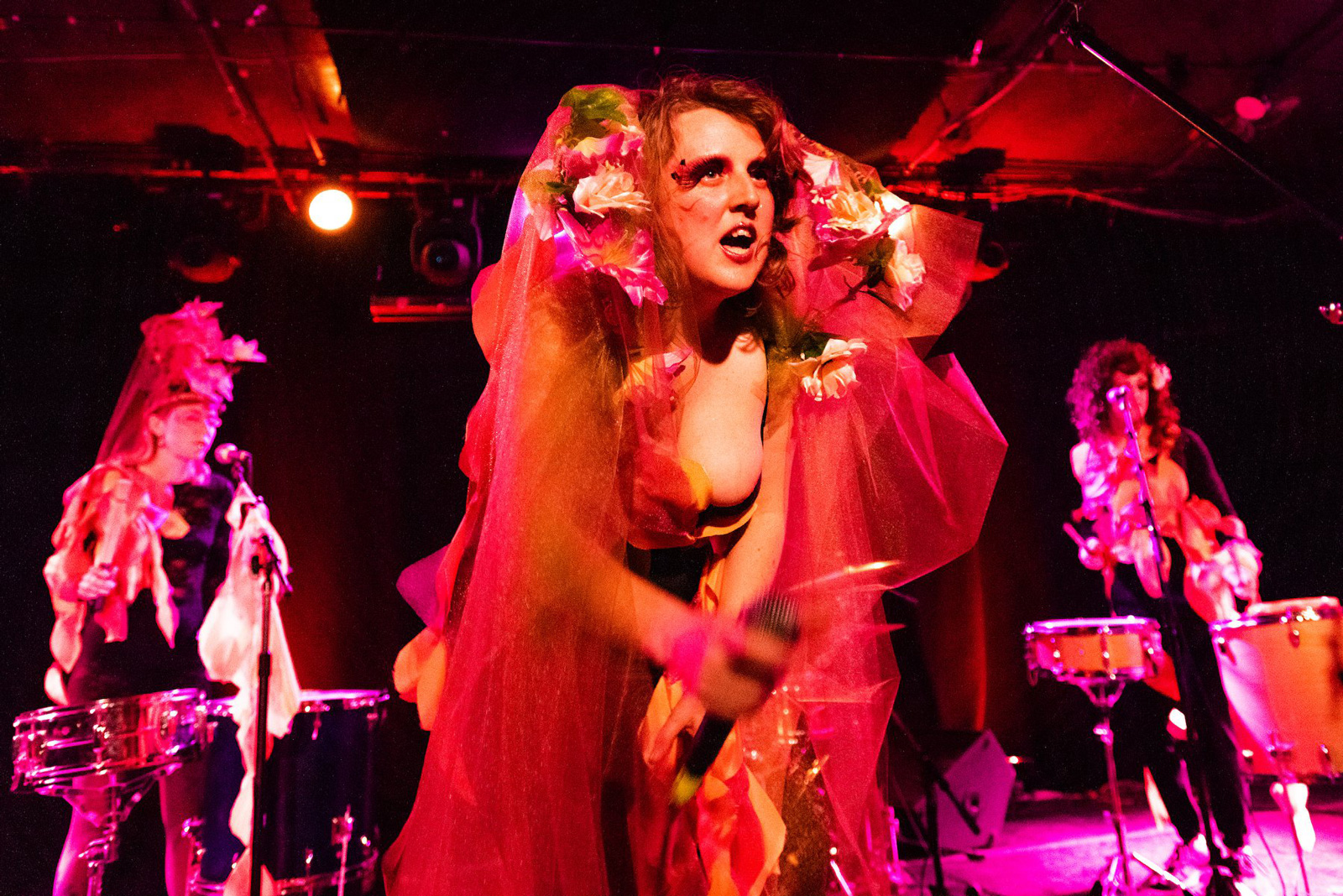 A photograph of Emily Bate performing in a show, clad in billowing red garments and bouquets of flowers, scintillating makeup and hair glowing red in the stage lights, a look of delighted fury upon her face. Bandmates are similarly costumed on the stage behind her.